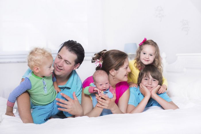 Big family with four kids relaxing in bed on a sunny morning. Parents with newborn baby, toddler boy, preschooler girl and teenager son in a white bedroom. Mother and father playing with children.