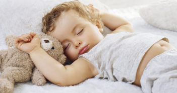 Sleeping baby boy arms raised up with a Teddy bear on a white blanket