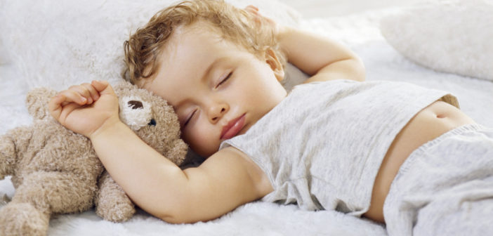 Sleeping baby boy arms raised up with a Teddy bear on a white blanket