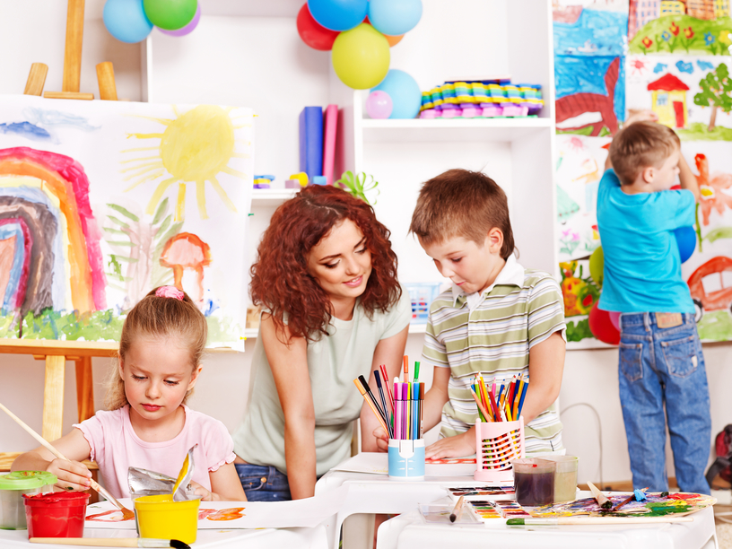 Children with teacher painting at easel in school.