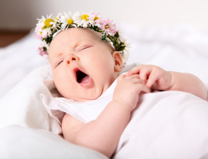 Portrait of a yawning baby girl on a light background with a wreath of flowers on her head